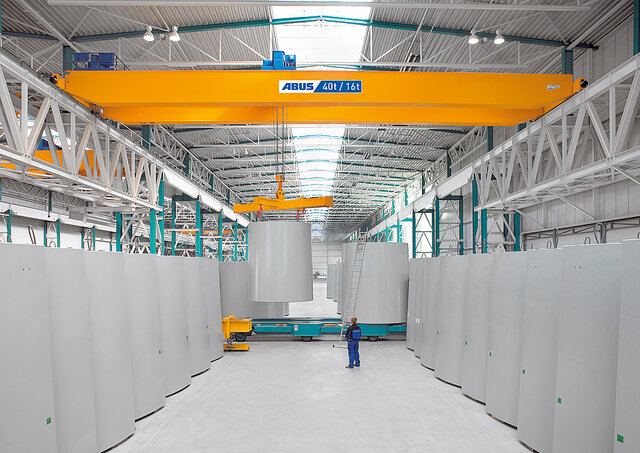 Una grúa puente se desplaza por una nave industrial transportando grandes cargas, mientras un trabajador se encuentra en la zona de trabajo.