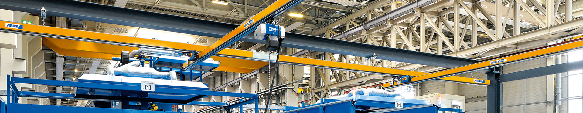 Em um galpão industrial, podem ser vistos vários trilhos suspensos de viga única do tipo EHB. Em primeiro plano, um trabalhador opera um guincho de corrente com um botão suspenso. O guincho de corrente se move com um carro deslizante no perfil da viga principal.