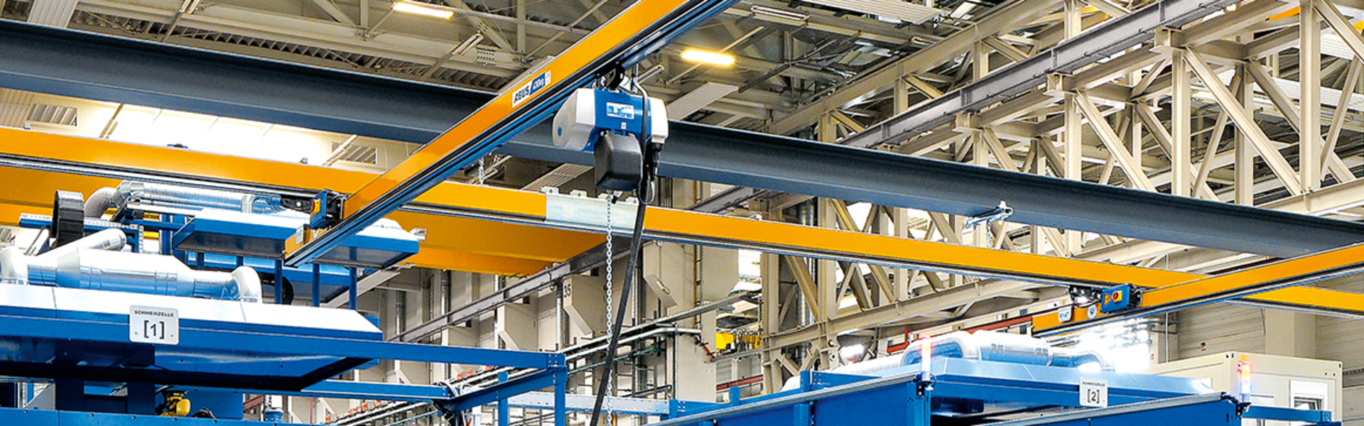 Em um galpão industrial, podem ser vistos vários trilhos suspensos de viga única do tipo EHB. Em primeiro plano, um trabalhador opera um guincho de corrente com um botão suspenso. O guincho de corrente se move com um carro deslizante no perfil da viga principal.