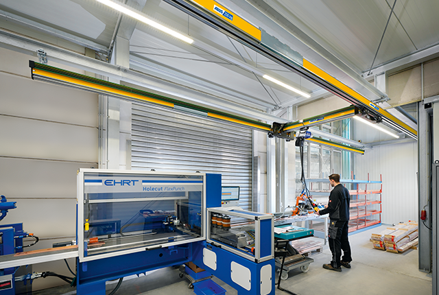 La grúa de una viga EHB-X desplaza una carga en una nave industrial, mientras un empleado trabaja en una máquina al fondo.